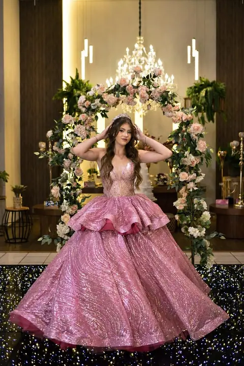Debutante de vestido rosa cintilante com as mãos no cabelo, diante de um arco de flores e lustre