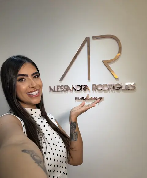 Mulher sorrindo e apontando para o letreiro Alessandra Rodrigues makeup na parede do espaço