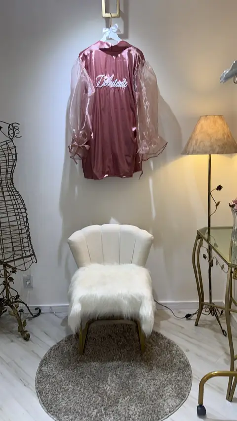 Robe rosa bordado com a palavra Debutante pendurado na parede, acima de uma poltrona com pelúcia branca