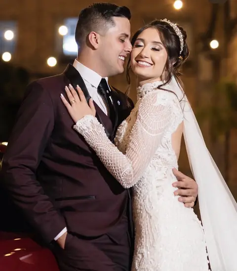 Noiva sorrindo de olhos fechados, com tiara e véu, abraçada ao noivo de terno vinho
