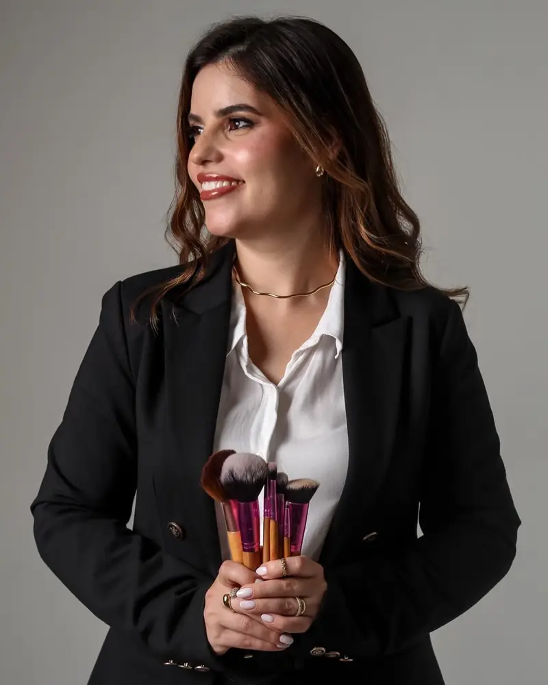 Alessandra Rodrigues de blazer preto e camisa branca, sorrindo e segurando seus pincéis de maquiagem