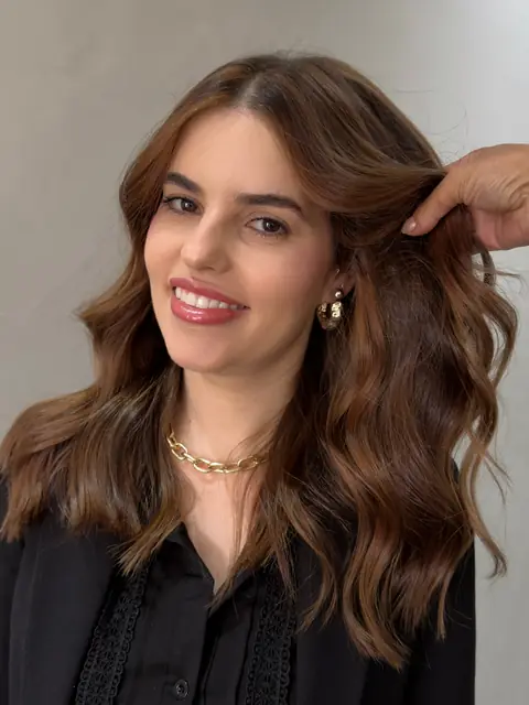 Mulher de cabelo castanho iluminado em ondas, com a mão ajeitando os fios