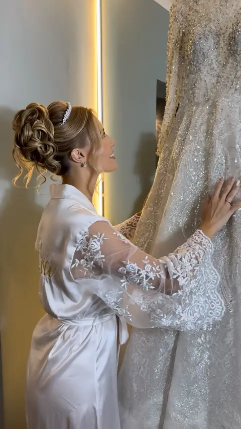 Noiva de robe branco com coque volumoso e tiara de pérolas, de perfil, tocando o vestido pendurado