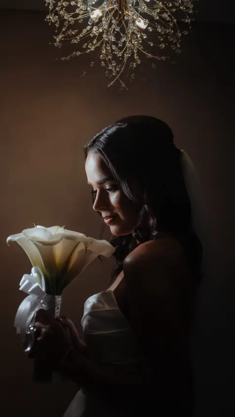 Noiva de perfil em luz baixa segurando buquê de copos-de-leite sob um lustre, em tons escuros e dourados