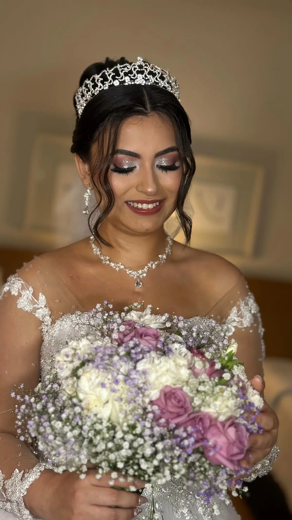 Noiva sorrindo com tiara e colar, segurando buquê lilás e rosa com mosquitinho