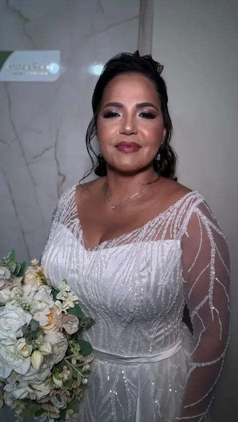 Noiva de vestido bordado com manga transparente e buquê branco, diante de parede de mármore