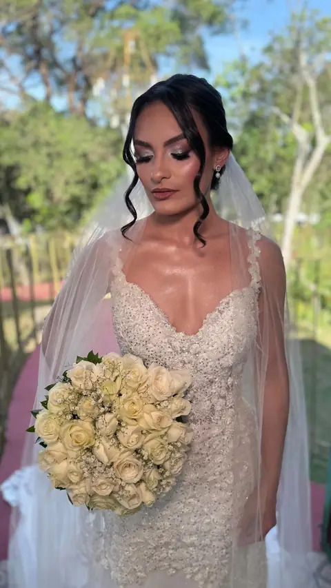 Noiva ao ar livre com véu, cabelo preso com mechas soltas e buquê de rosas brancas