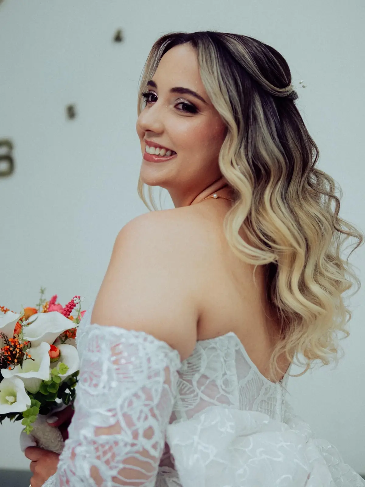 Noiva loira olhando por cima do ombro, com cabelo semipreso ondulado e vestido de renda com as costas à mostra