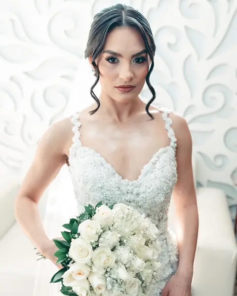 Noiva olhando para a câmera, com cabelo preso e mechas soltas, vestido bordado e buquê de rosas brancas