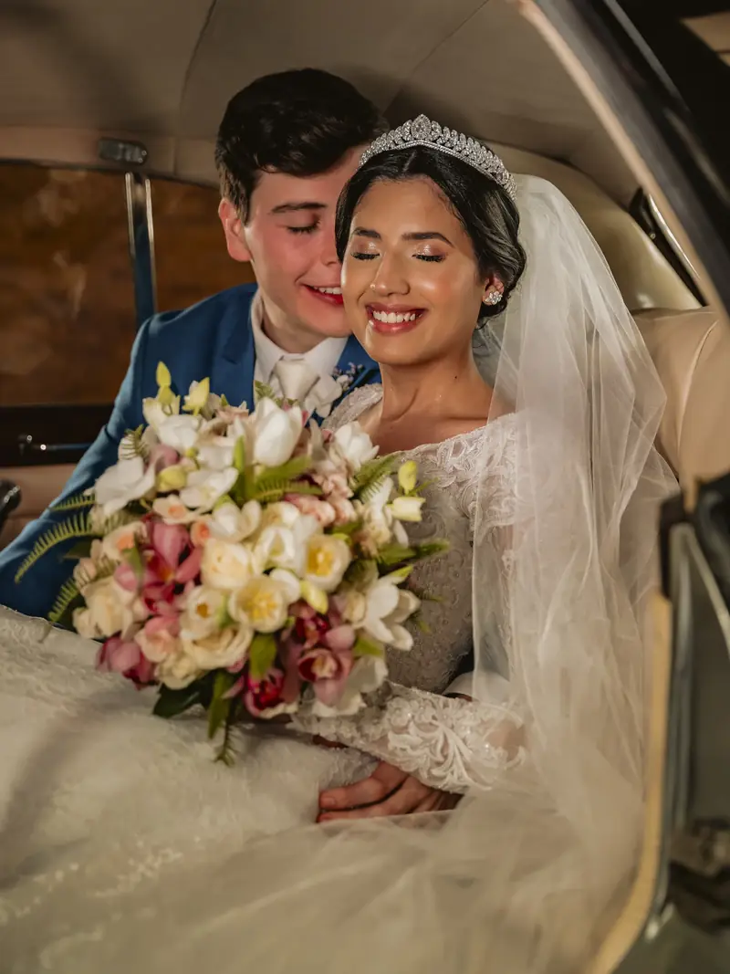 Casal de noivos no carro, noiva de tiara sorrindo de olhos fechados com buquê colorido
