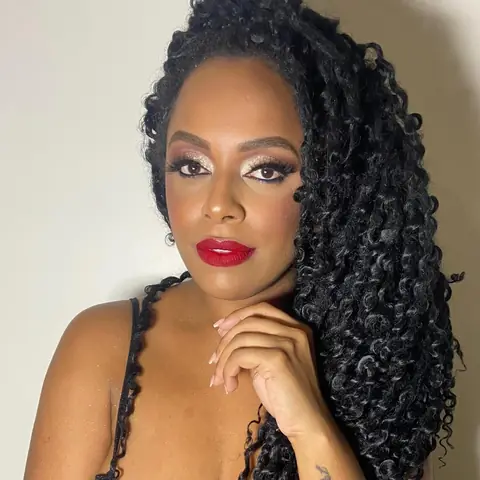 Mulher de cabelo cacheado volumoso com batom vermelho e olho iluminado, com a mão no queixo