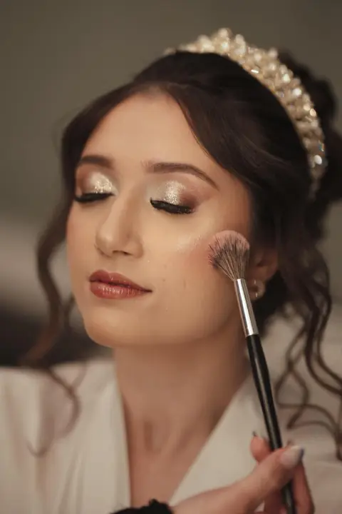 Noiva de tiara e olhos fechados recebendo blush com pincel, com sombra dourada iluminada