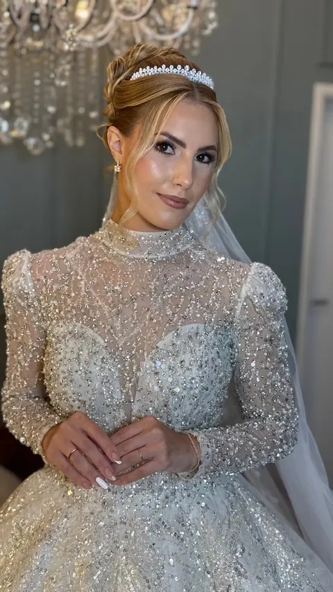Noiva loira com coque, tiara e vestido bordado de brilho com gola alta, sob lustre de cristal